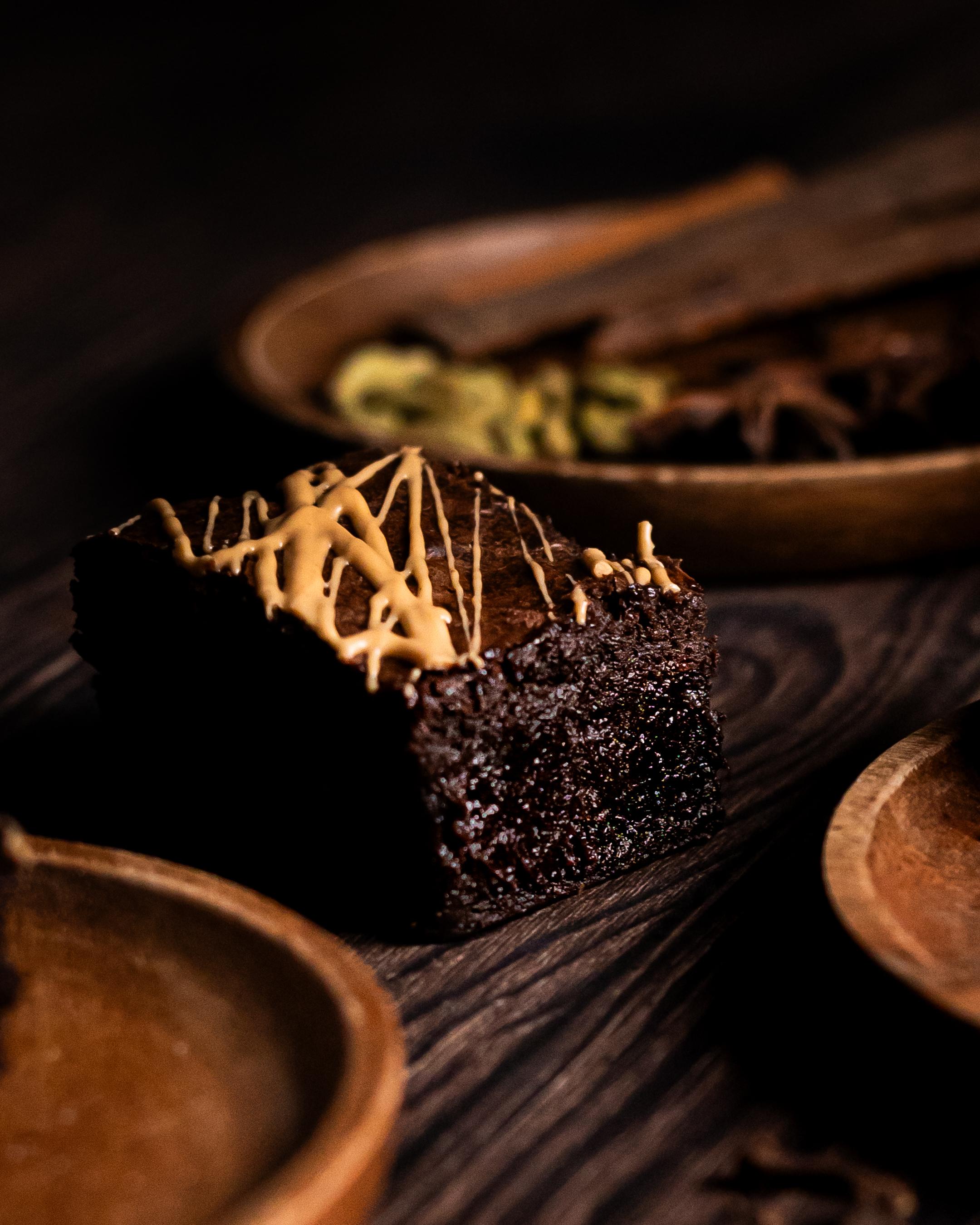a single masala chai brownie topped with gold chocolate. A wooden plate of aromatic spices in the background