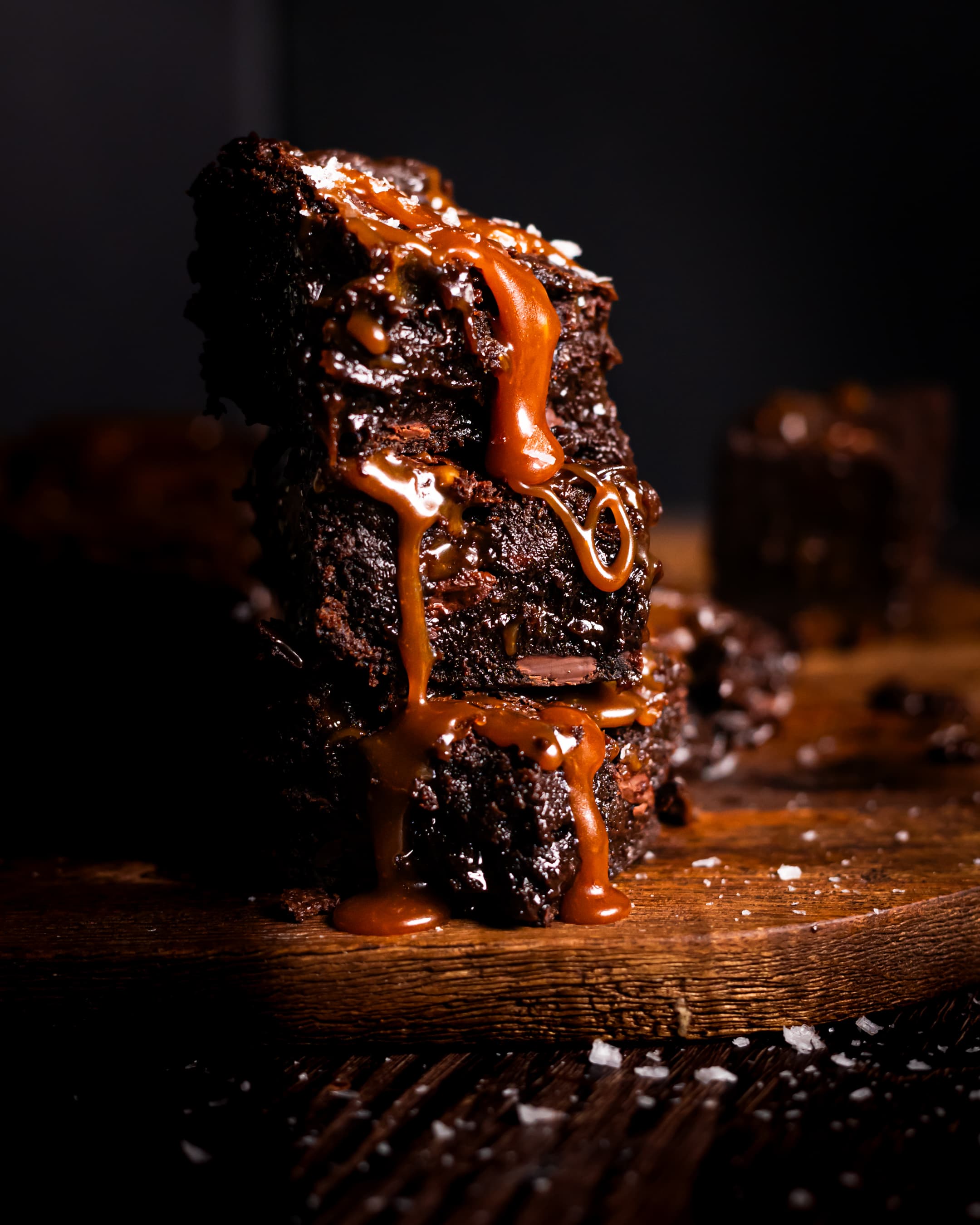 A stack of 3 salted caramel brownies with salted caramel dripping down, sprinkled with sea salt. Salted caramel brownies in the background and sea salt sprinkled in the foreground