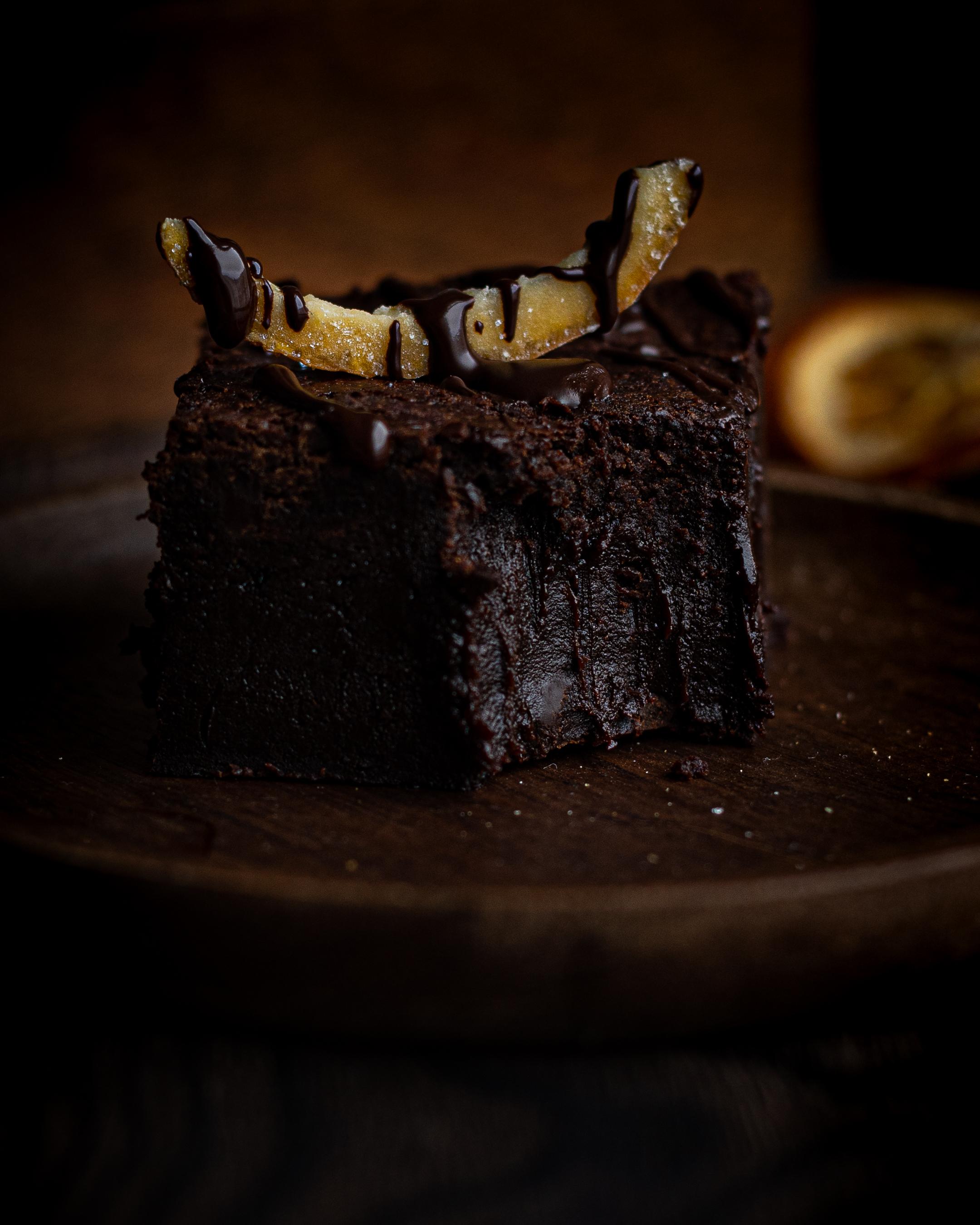a single chocolate orange brownie with a bite taken out, teethmarks showing in the gooey centre of the brownie. Topped with a candied orange crown. Dried oranges in background