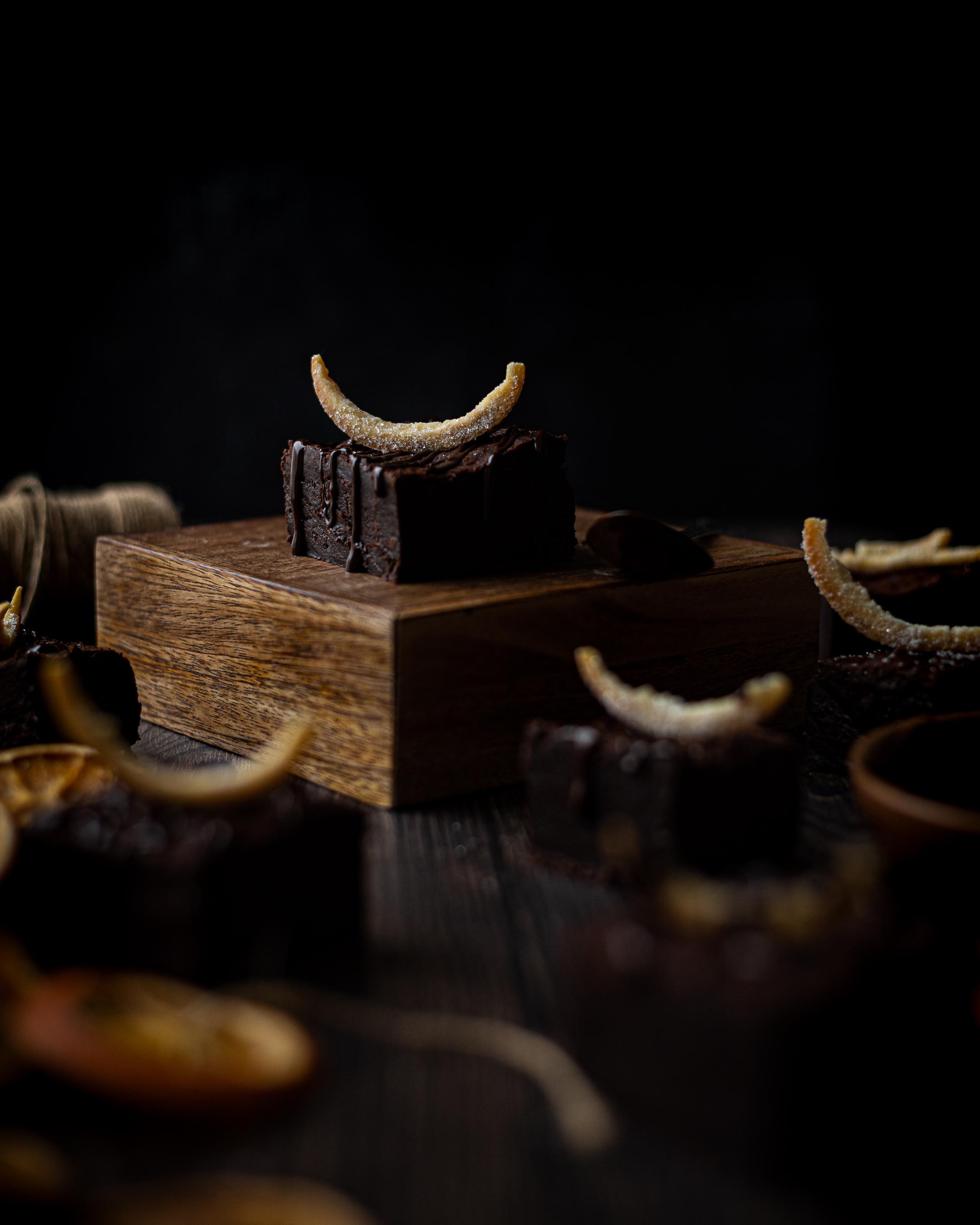 a chocolate orange brownie on a wooden platform, topped with a candied orange crown, surrounded by other chocolate orange brownies and dried oranges in the foreground